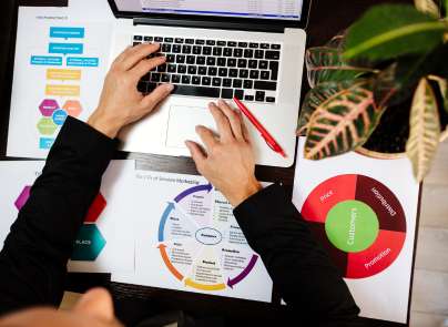 Hände am Laptop mit Papieren, darauf Statistiken und Diagramme zur Produktrecherche
